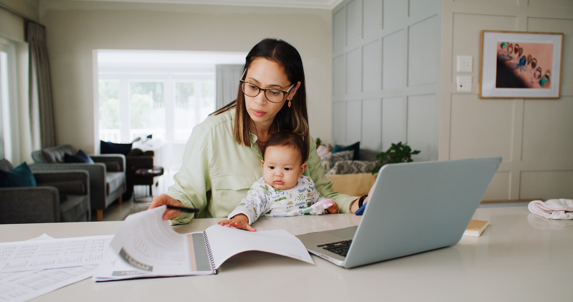 Woman wearing glasses sits at a kitchen island with a baby on her lap, looking at papers beside an open laptop.